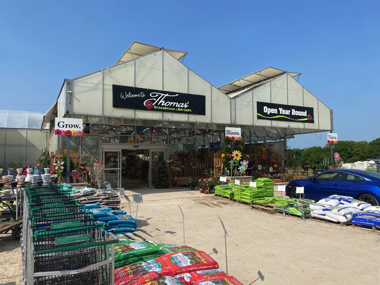 Exterior view of Thomas Greenhouse & Gardens in Mukwonago, Wisconsin, showing entrance, garden supplies, and seasonal decor.